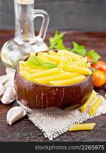 Wheat flour penne pasta in a bowl of coconut shells on sacking, tomatoes, garlic, vegetable oil in a decanter and parsley on wooden board background