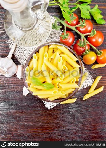 Wheat flour penne paste in a bowl of coconut shells on sacking, tomatoes, garlic, vegetable oil in a glass decanter and parsley on a wooden board background from above