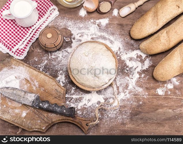 wheat yeast dough for bread and rolls in a wooden bowl on a table in the middle of the ingredients, top view