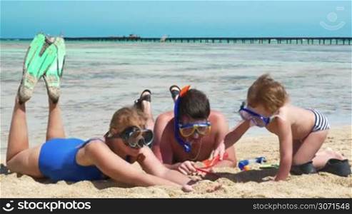 While resting after the snorkel diving, parents and son are playing in sand on the beach.