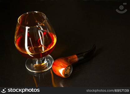 Whiskey glass and smoking pipe on black background. Cognac glass. Brandy glassful. Cognac france. Scotch drink. Smoking pipe.. Whiskey glass and smoking pipe on black background
