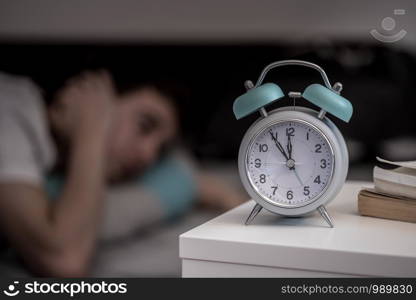 White alarm clock on the bedside table. Young tries to sleep in the background, insomnia
