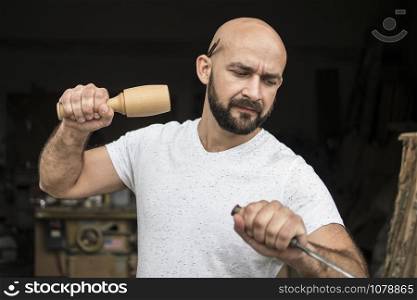 white bald carpenter with beard in white t-shirt works as a chisel and a mallet