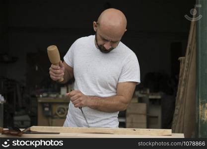 white bald carpenter with beard in white t-shirt works as a chisel and a mallet