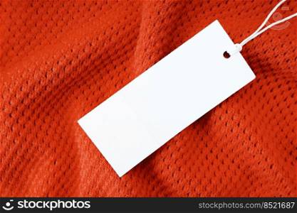 White blank rectangular clothing tag, label mockup template on red knitted fabric background . Price tag label with copy space for text. Shopping, sale, price, fashion, discount concept. White blank rectangular clothing tag on red knit fabric background. Shopping, sale, discount mockup