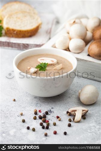 White bowl plate of creamy chestnut champignon mushroom soup on light kitchen background and box of raw mushrooms and fresh bread.