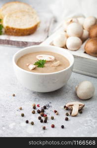 White bowl plate of creamy chestnut champignon mushroom soup on light kitchen background and box of raw mushrooms and fresh bread.