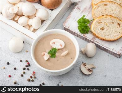 White bowl plate of creamy chestnut champignon mushroom soup on light kitchen background and box of raw mushrooms and fresh bread. Top view