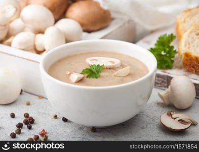 White bowl plate of creamy chestnut champignon mushroom soup on light kitchen background and box of raw mushrooms and fresh bread.