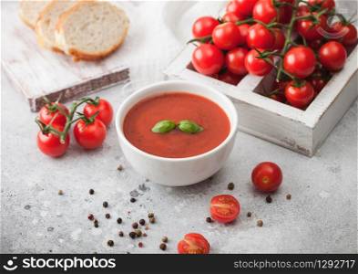 White bowl plate of creamy tomato soup with pepper on light background with box of raw tomatoes and bread.