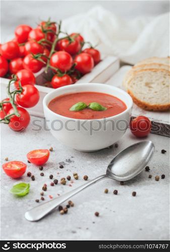 White bowl plate of creamy tomato soup with spoon on light background with box of raw tomatoes and bread.