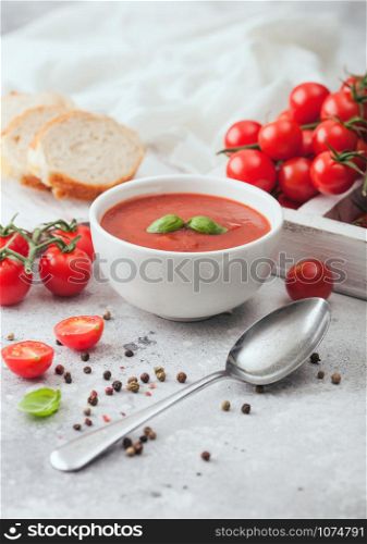 White bowl plate of creamy tomato soup with spoon on light background with box of raw tomatoes and bread.