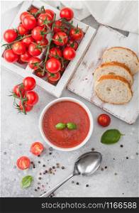 White bowl plate of creamy tomato soup with spoon on light background with box of raw tomatoes and bread. Top view