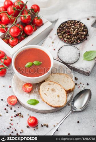 White bowl plate of creamy tomato soup with spoon on light background with box of raw tomatoes and bread. Top view