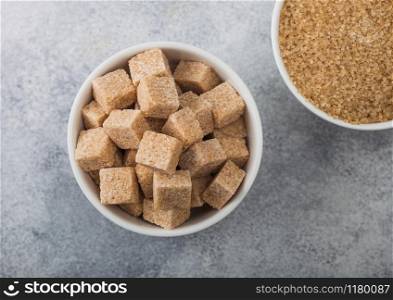 White bowl plates of natural brown sugar cubes and refined sugar on light background. Top view