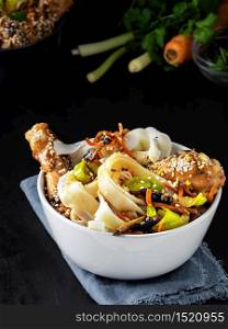 White bowl with noodles is located on a dark background. Asian-style lunch, noodles with chicken in teriyaki sauce, vegetables, spices and microgreens, ingredients for noodles on the table. Noodles with vegetables, chicken and teriyaki sauce, closeup.