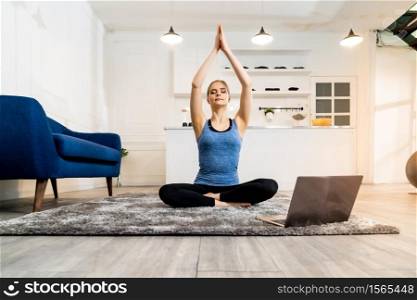 White caucasian fit sporty woman doing yoga Sukhasana sit pose in living room at home watching online tutorials from trainer on laptop. Working out, fitness sport and healthy lifestyle concept.