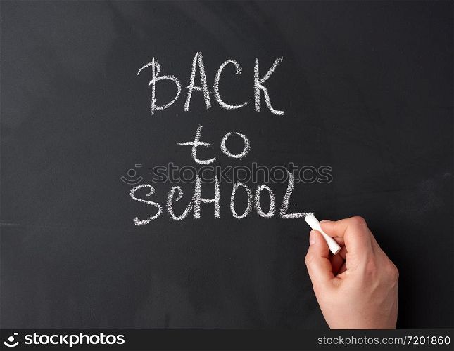 white chalk back to school on black chalkboard, hand holds a piece of white chalk, close up
