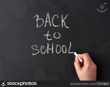 white chalk back to school on black chalkboard, hand holds a piece of white chalk, close up