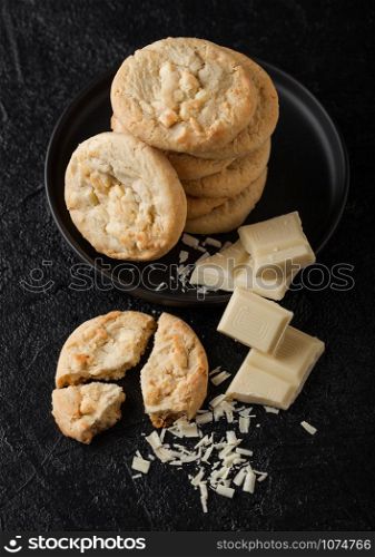 White chocolate biscuit cookies on black ceramic plate with chocolate blocks and curls on dark table background.