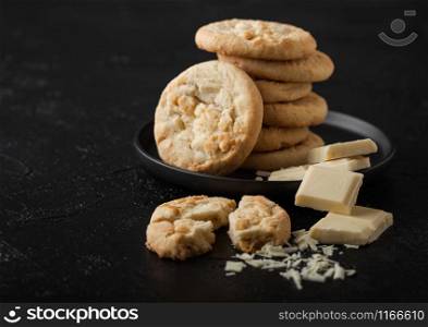 White chocolate biscuit cookies on black ceramic plate with chocolate blocks and curls on dark table background.