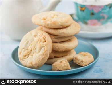 White chocolate biscuit cookies on blue ceramic plate with tea pot and cup on blue table background.