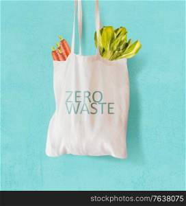 White cloth bag with inscription &rsquo;zero waste&rsquo; filled with vegetables hanging in front of bright blue background. Ecological, sustainable concept. Front view