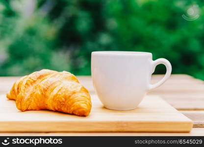 White Coffee Cup And French Croissant On Wooden Table In Green Garden