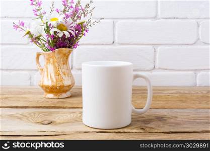 White coffee mug mockup with chamomile and purple flowers in gol. White coffee mug mockup with white chamomile and purple field flowers in golden pitcher vase. Empty mug mock up for design promotion.