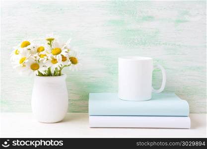 White coffee mug mockup with white field chamomile bouquet in ha. White coffee mug mockup with white field chamomile bouquet in handmade rustic vase and books. Empty mug mock up for design promotion.