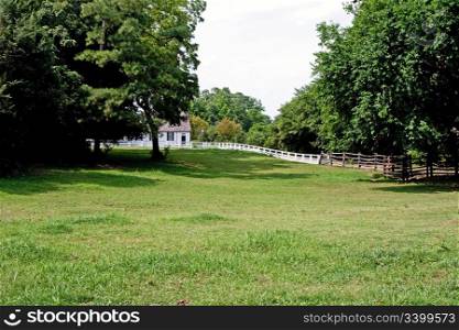 White colonial wooden farm house with a meadow grass field and trees in front barricaded with a white fence.