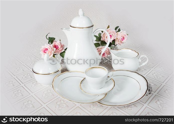 White crockery for coffee: coffeepot, cup, serving plate and sugar bowl as well as rose flowers are on a beautiful tablecloths