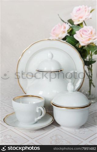 White crockery for tea: teapot, cup, serving plate and sugar bowl as well as rose on a beautiful tablecloths