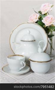 White crockery for tea: teapot, cup, serving plate and sugar bowl as well as rose on a beautiful tablecloths