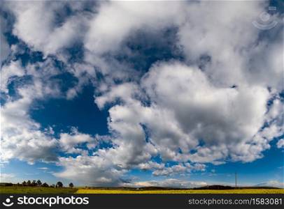 White cumulus clouds in blue sky over spring rapeseed evening fields and rural hills high resolution background. Natural seasonal, weather, climate, countryside beauty concept high resolution scene.