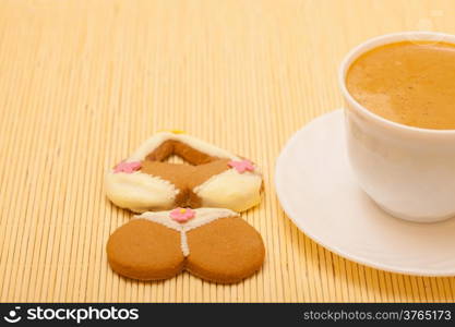 White cup of coffe with funny bikini underwear shape gingerbread cake cookie sweet dessert with yellow icing and pink decoration on beige bamboo mat background