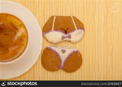 White cup of coffe with funny bikini underwear shape gingerbread cake cookie sweet dessert with white icing and violet decoration on beige bamboo mat