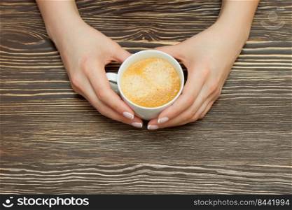 White cup of coffee in female hands on brown wooden table. Top view, flat lay with copy space for text. White cup of coffee in female hand