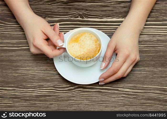 White cup of coffee in female hands on brown wooden table. Top view, flat lay with copy space for text. White cup of coffee in female hand