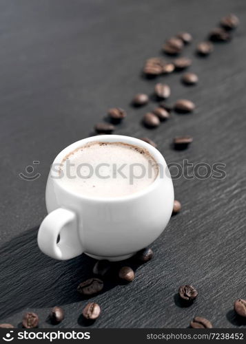 White cup of coffee with milk on a black stone background. Roasted coffee beans are located around a cup of coffee.