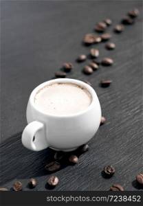 White cup of coffee with milk on a black stone background. Roasted coffee beans are located around a cup of coffee.