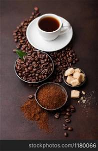 White cup of fresh raw organic coffee with beans and ground powder with cane sugar cubes with coffee tree leaf on dark background. Top view
