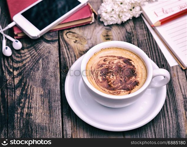 white cup with cappuccino behind the mobile phone with headphones on a gray wooden background