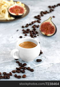 White cup with freshly brewed espresso on a gray background. Roasted coffee beans are located around a cup of coffee. In the background is fresh slaced figs and homemade cheese.