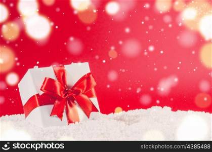 white decorative christmas gift box with ribbon on snow against red festive background. christmas gift box