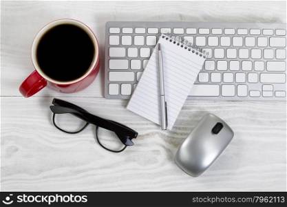 White desktop with red coffee cup, partial keyboard, mouse, reading glasses, paper and pen.