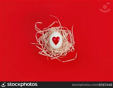 White egg with heart shape in decorative nest on a red background.. White egg with heart shape in decorative nest on red background.
