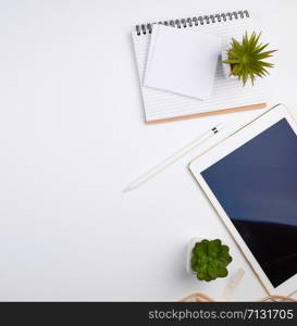 white electronic tablet with a blank screen and a pencil, near pots with green plants on a white table, top view, designer workplace