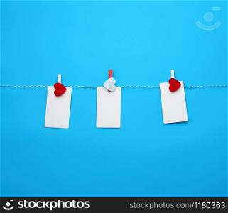 white empty paper rectangular business cards on decorative clothespins with a red heart hanging on a rope, blue background, place for an inscription