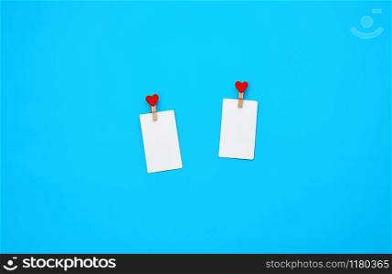 white empty paper rectangular business cards on decorative clothespins with a red heart lie on a blue background, place for an inscription